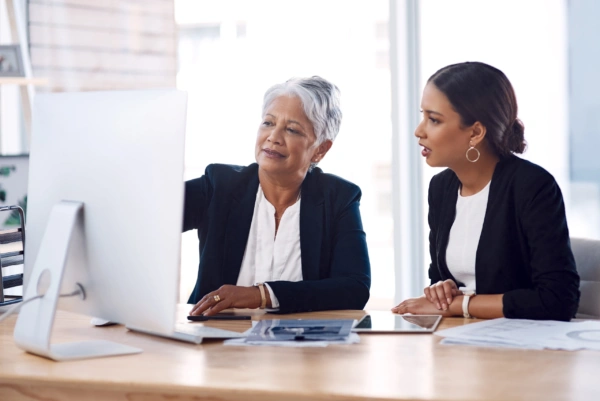 Two women in business attire sit at a desk, looking at a computer screen. One woman points at the monitor while the other listens attentively. Papers and a tablet are on the desk in front of them.