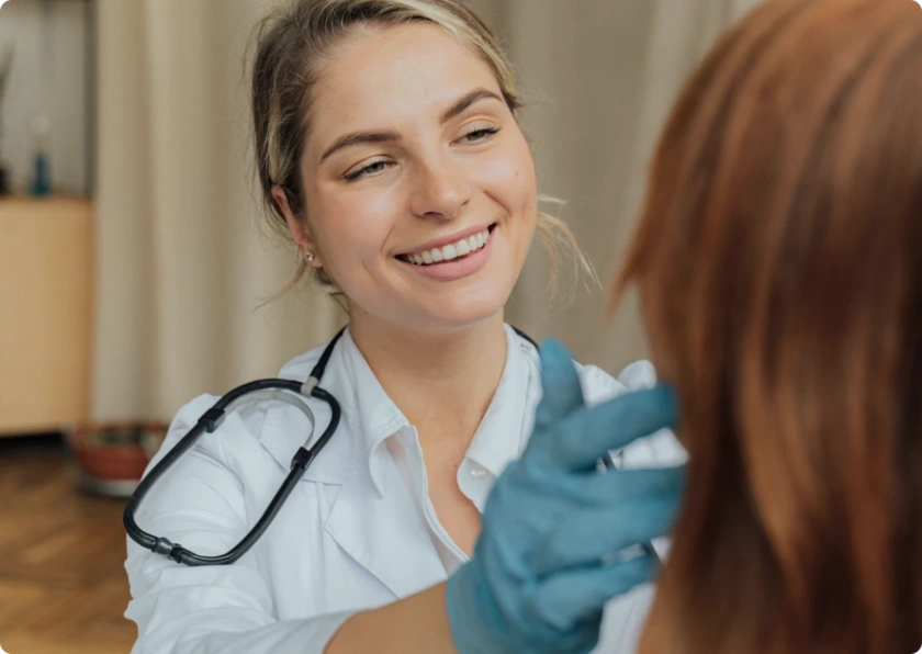 A smiling healthcare professional with a stethoscope around her neck and blue gloves demonstrates her professional skills while examining a patient, whose back and brown hair are visible in the foreground.