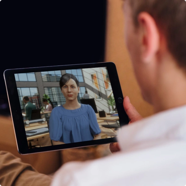 A person holds a tablet showing an On-Demand Simulation Training video call with a woman in a blue blouse, sitting in a bright office with large windows and people working in the background.