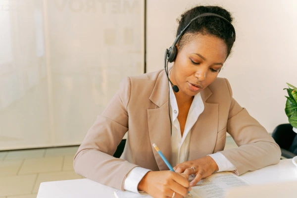 A woman wearing a headset and beige blazer sits at a desk, writing notes on a document with a pencil while talking. A potted plant is on the desk beside her.