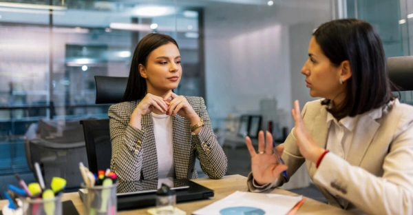 Two women sit across from each other at an office desk, engaged in a serious conversation as part of training simulations. One woman listens intently while the other gestures with her hands. Office supplies and documents are visible on the desk.