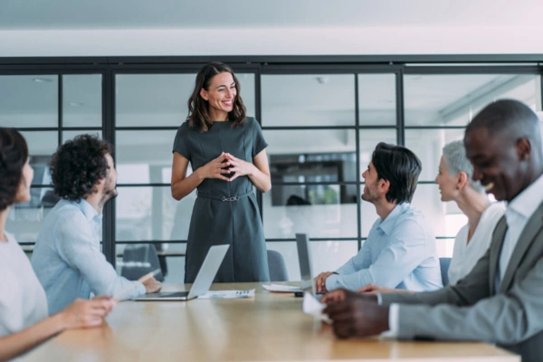 A woman stands and smiles while speaking to a group of five colleagues seated around a conference table in a modern office, leading an engaging discussion on leadership development as the team listens attentively with laptops and notepads open.