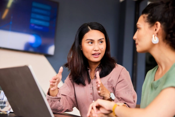 Two women sit at a table with a laptop, engaged in conversation about leadership development. One gestures while speaking passionately, and the other listens attentively. A blurred screen is visible in the background.
