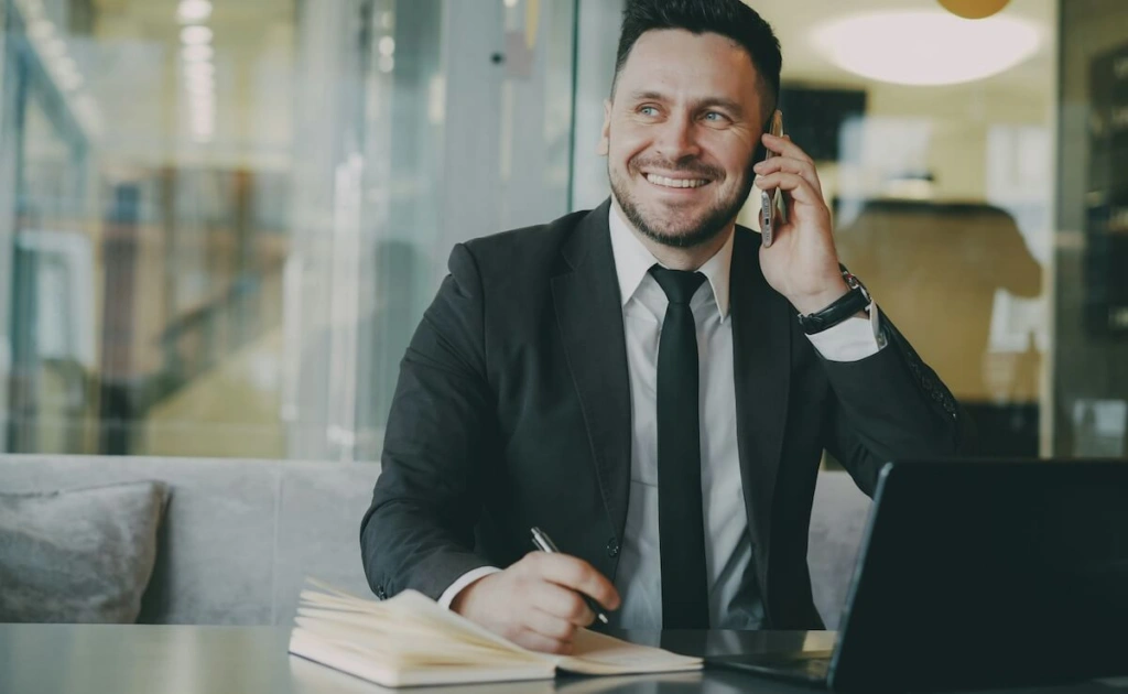 A smiling man in a dark suit talks on his phone while sitting at a desk with an open laptop and notebook, holding a pen, in a modern office setting.