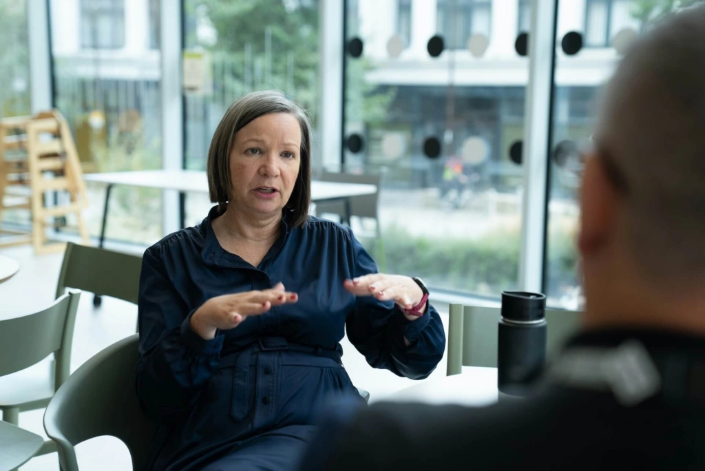 A woman with shoulder-length hair, wearing a dark blue blouse, sits at a table gesturing with her hands while discussing managing team conflict with an unseen person in a modern, glass-walled indoor setting.